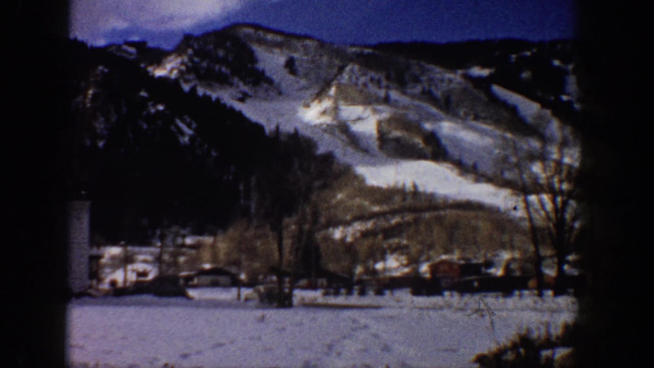 Wide view of a rocky snow-covered mountain range above a valley with small structures and parked vehicles, shot on 8mm film.