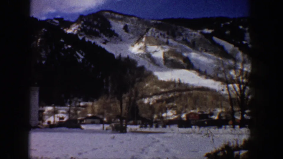 8mm home movie frame of a snow-dusted mountain range above a small valley settlement with parked vehicles, possibly Aspen, Colorado, circa 1961.
