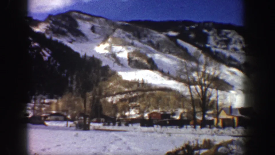 8mm home movie frame showing a snow-dusted mountain, winter valley, and a small wooden cabin at frame edge, possibly Aspen, Colorado, circa 1961.
