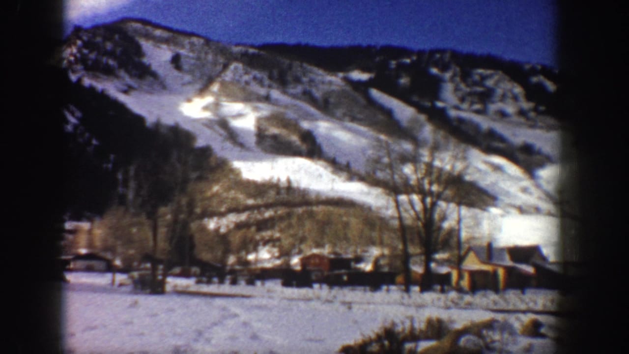 8mm film frame, Aspen, Colorado, c.1961 — steep snow-covered mountain above rustic wooden buildings, bare winter trees, and a flat snow-covered valley floor.