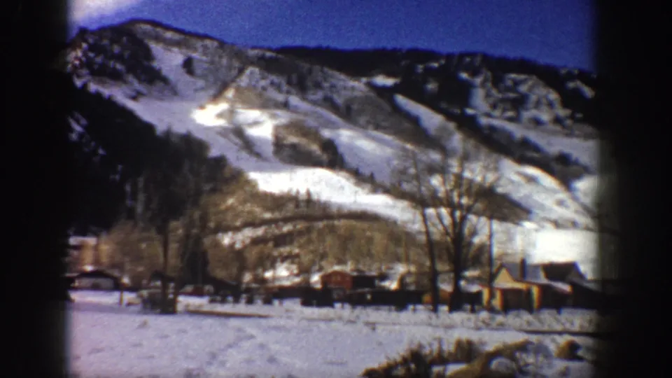8mm film frame, Aspen, Colorado, c.1961 — steep snow-covered mountain above rustic wooden buildings, bare winter trees, and a flat snow-covered valley floor.