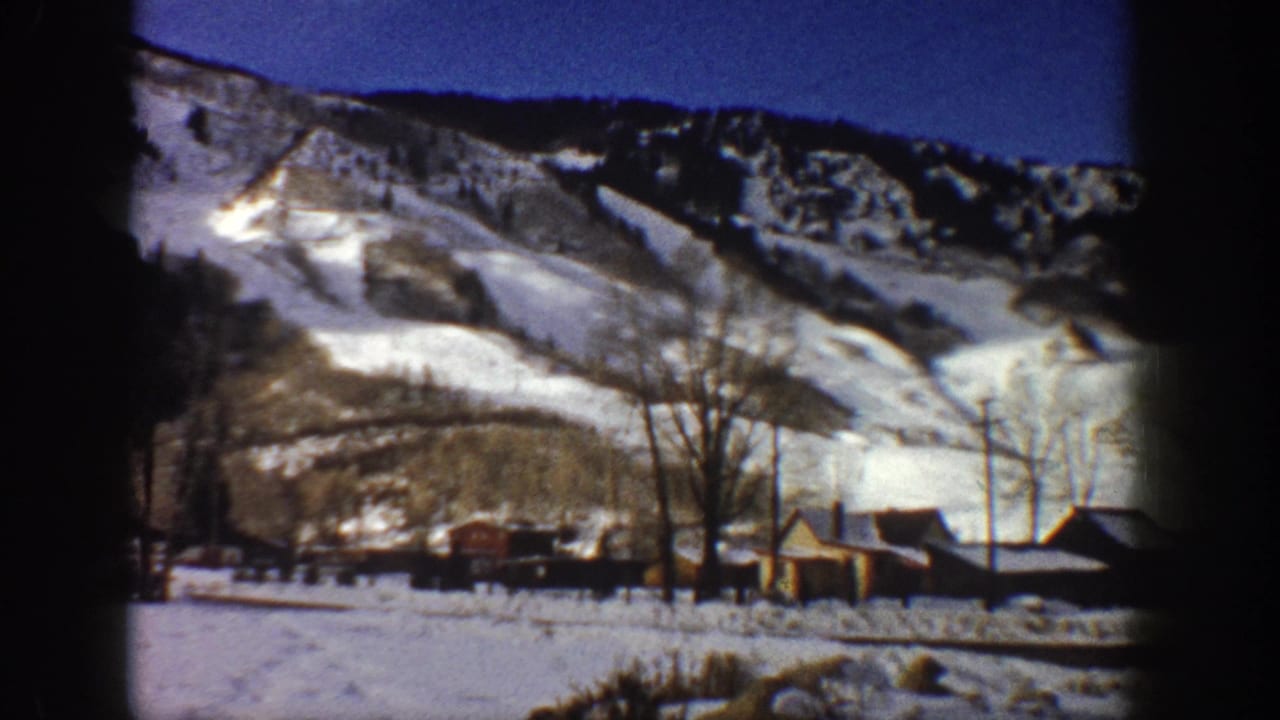 8mm pan frame, Aspen, Colorado, c.1961 — snow-covered mountain slopes and a small settlement of wooden buildings at the valley floor, bare trees in the middle ground.