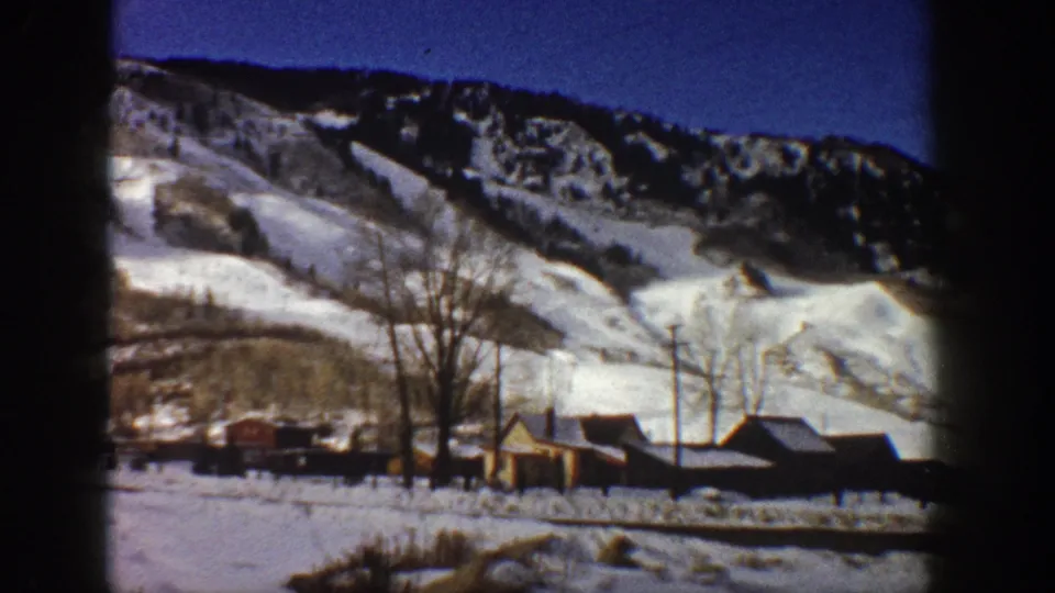 8mm frame, Aspen, Colorado, c.1961 — valley pan revealing additional rustic buildings, utility poles, and bare trees against snow-covered mountain and deep blue winter sky.