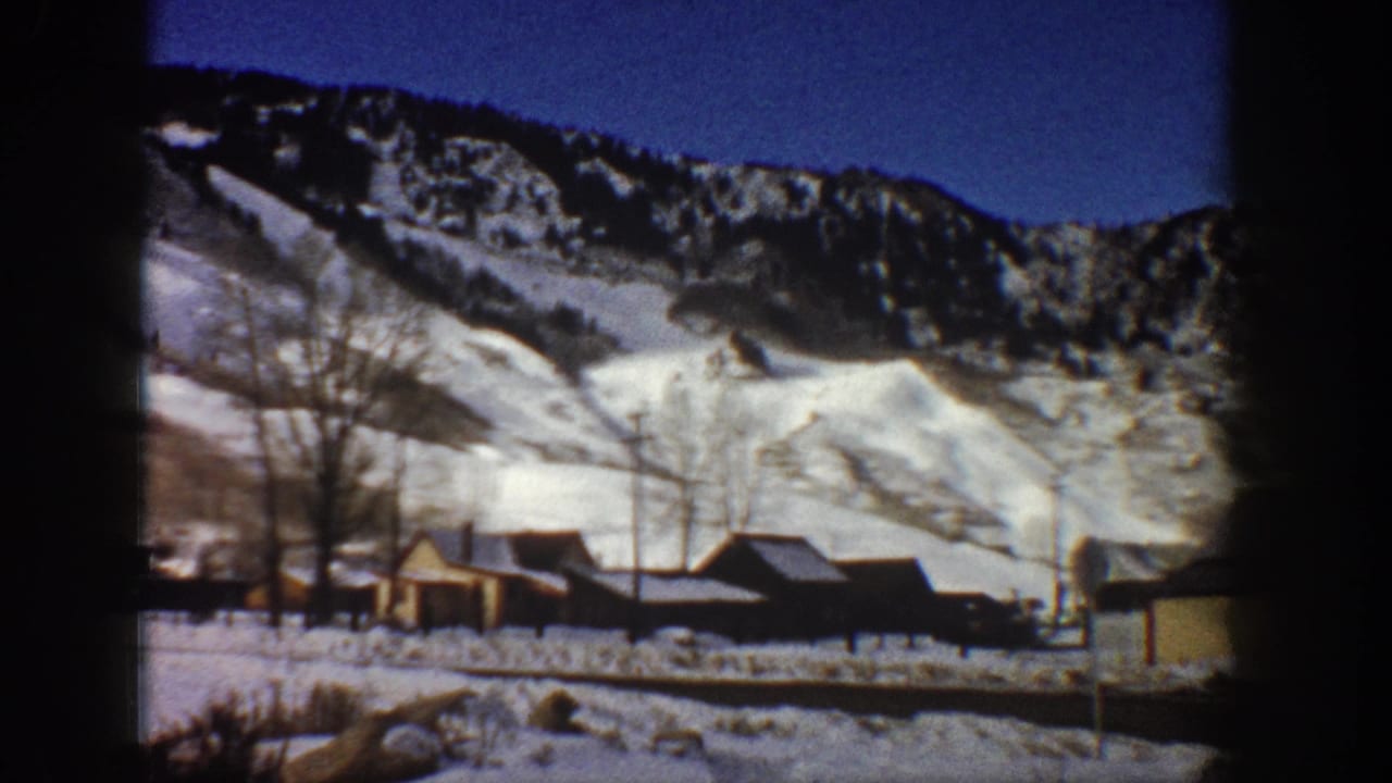 Rustic wooden buildings and bare trees below a snow-covered mountain ridge, shot on 8mm color film with dark edge vignetting.