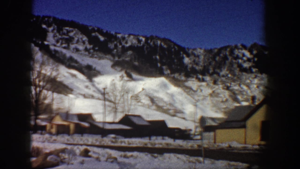 Pan continues across wooden buildings and bare trees below a snow-covered mountain, Aspen, Colorado, circa 1961, 8mm color footage.