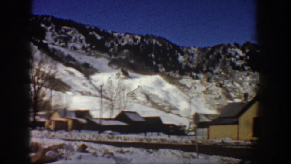 Pan continues across wooden buildings and bare trees below a snow-covered mountain, Aspen, Colorado, circa 1961, 8mm color footage.