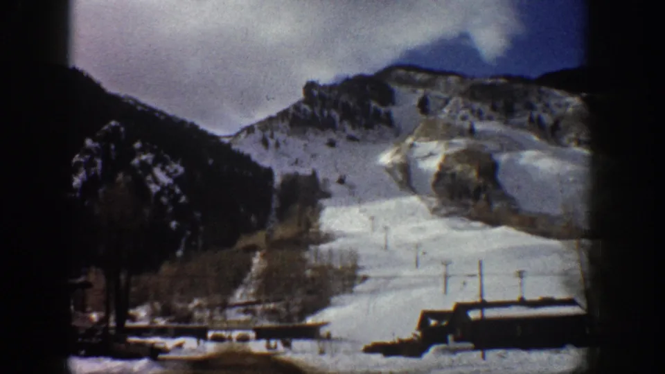 Snow-covered Rocky Mountain ridge with mist near the summit and low structures at lower right, Aspen, Colorado area, circa 1961, 8mm color.