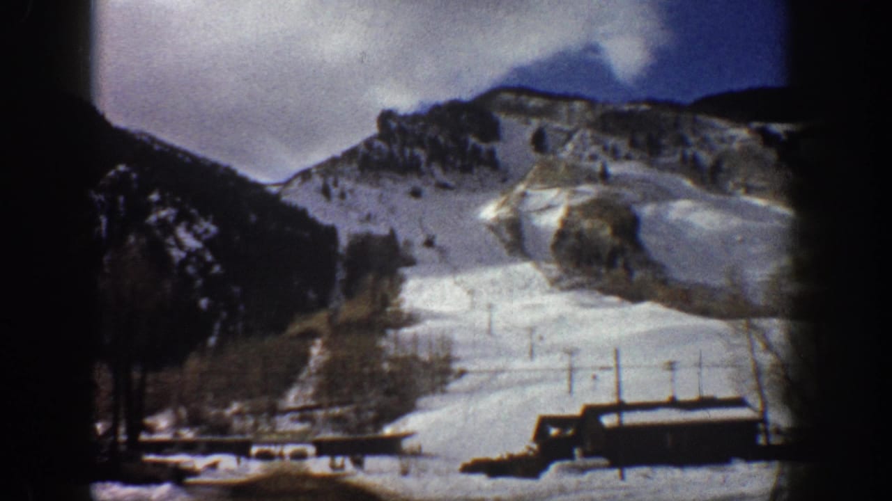 8mm home movie frame from Aspen, Colorado, circa 1961, showing a snow-covered ski slope, base lodge, and chairlift poles under a purple-gray overcast sky.