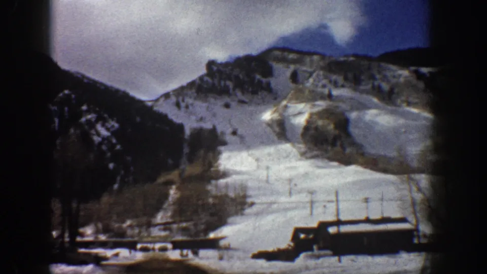 8mm home movie frame from Aspen, Colorado, circa 1961, showing a snow-covered ski slope, base lodge, and chairlift poles under a purple-gray overcast sky.