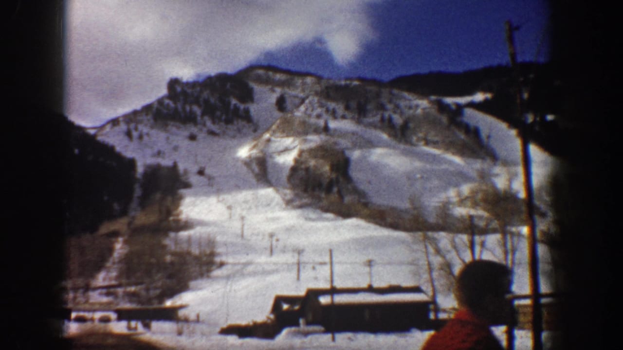 8mm home movie frame from Aspen, Colorado, circa 1961; snow-covered mountain slope, base lodge, chairlift poles, and a partially visible person in a red shirt at frame right.