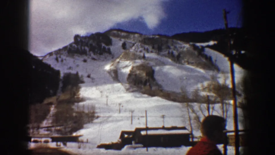 8mm home movie frame from Aspen, Colorado, circa 1961; snow-covered mountain slope, base lodge, chairlift poles, and a partially visible person in a red shirt at frame right.