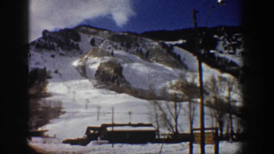 8mm home movie frame from Aspen, Colorado, circa 1961; snow-covered ski slope and base lodge under a clear blue sky with bare trees visible in the foreground.