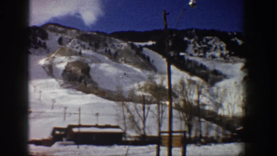 8mm home movie frame from Aspen, Colorado, circa 1961; snow-covered mountain and ski base area framed by heavy dark vignette borders under a partly cloudy blue sky.
