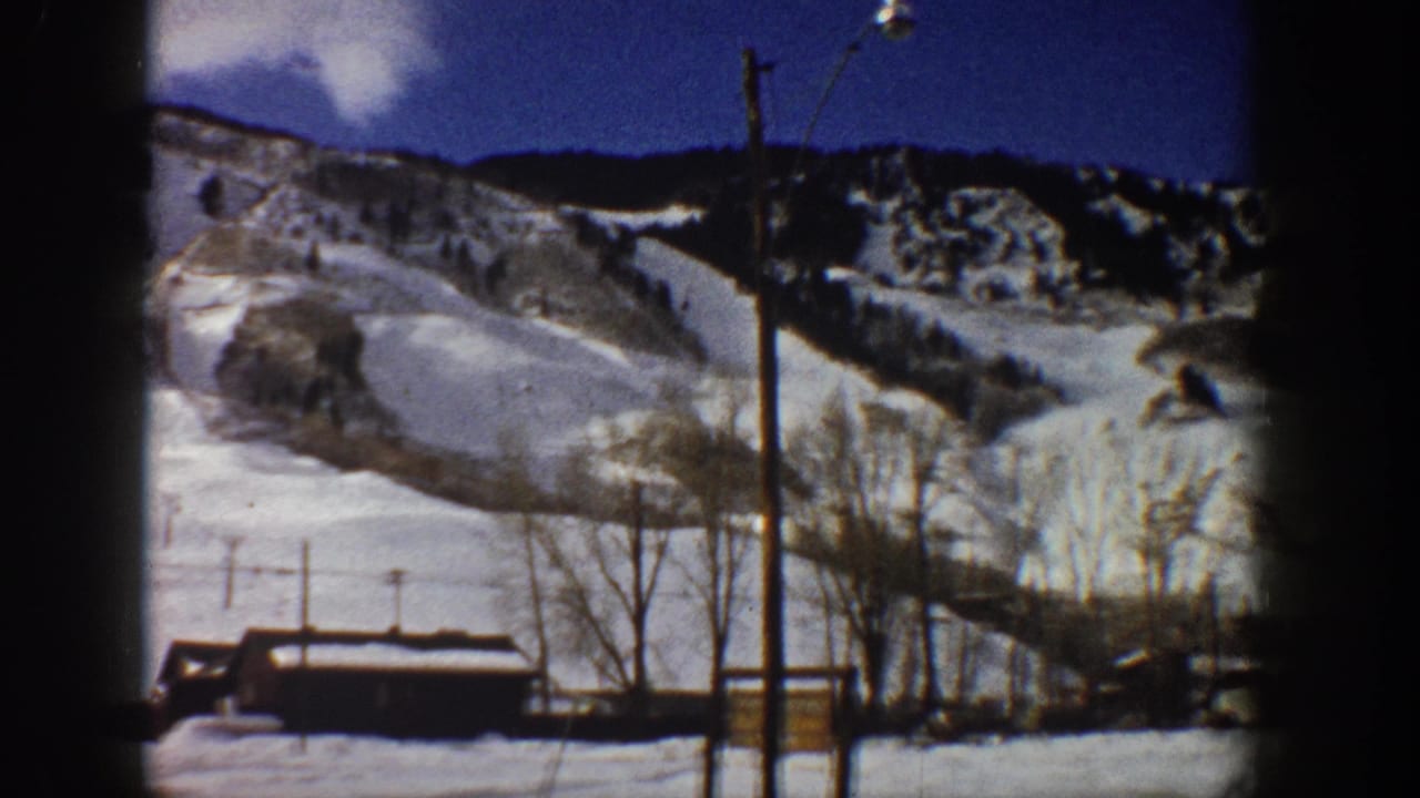 8mm home movie frame, Aspen, Colorado, c.1961 — snow-covered mountain slopes, bare aspen trees, a small wooden structure, and utility poles against a deep blue winter sky.