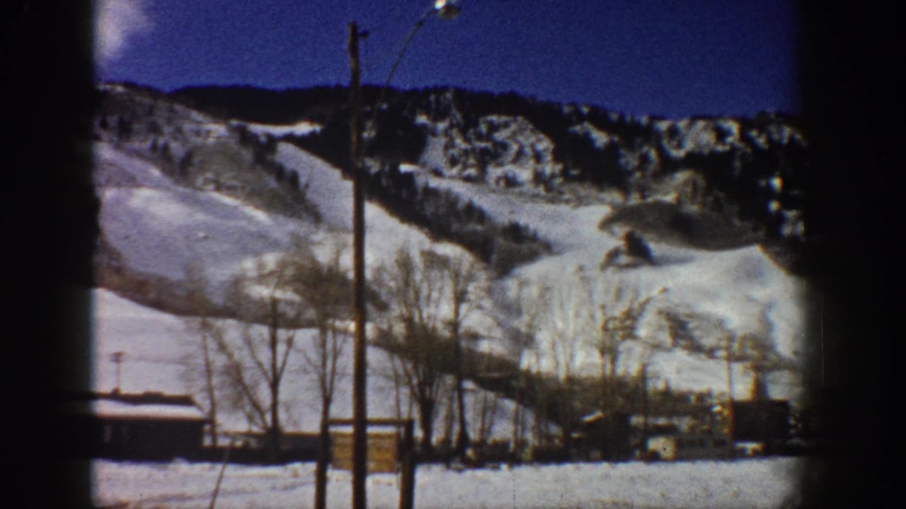 Pan continues over snow-covered Rocky Mountain slopes; bare winter trees and a low wooden building in foreground with utility pole.