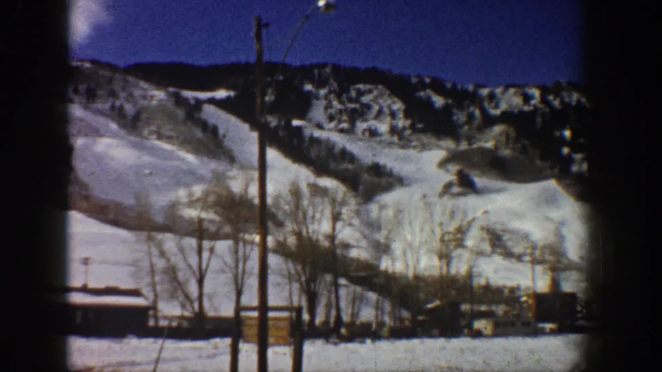 Mid-pan 8mm frame, Aspen, Colorado, c.1961 — snow-covered mountain slopes, bare winter trees, small building, and utility poles under deep blue sky.