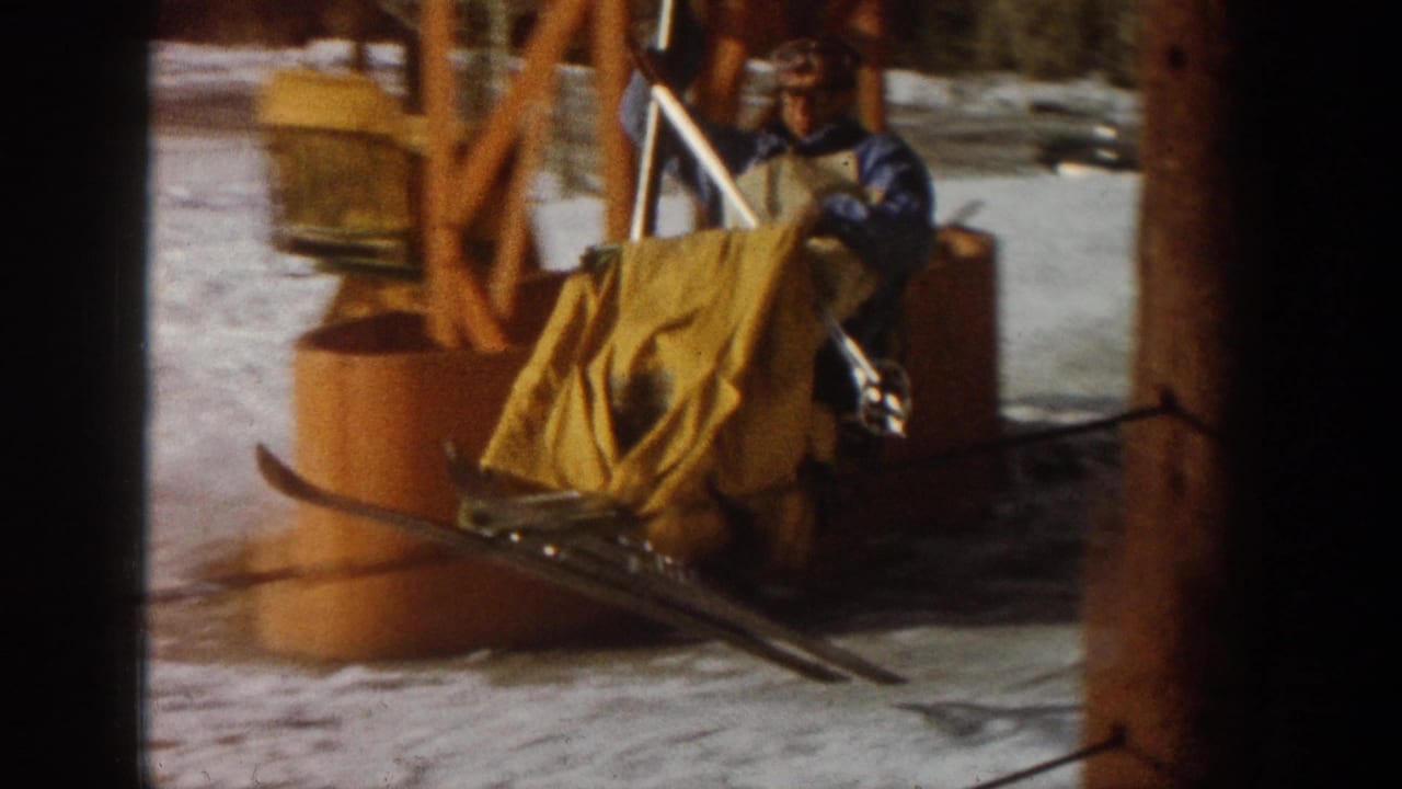 Man in blue ski jacket at chair lift loading area with yellow apron and orange metal lift frame, Aspen area circa 1961, 8mm footage.