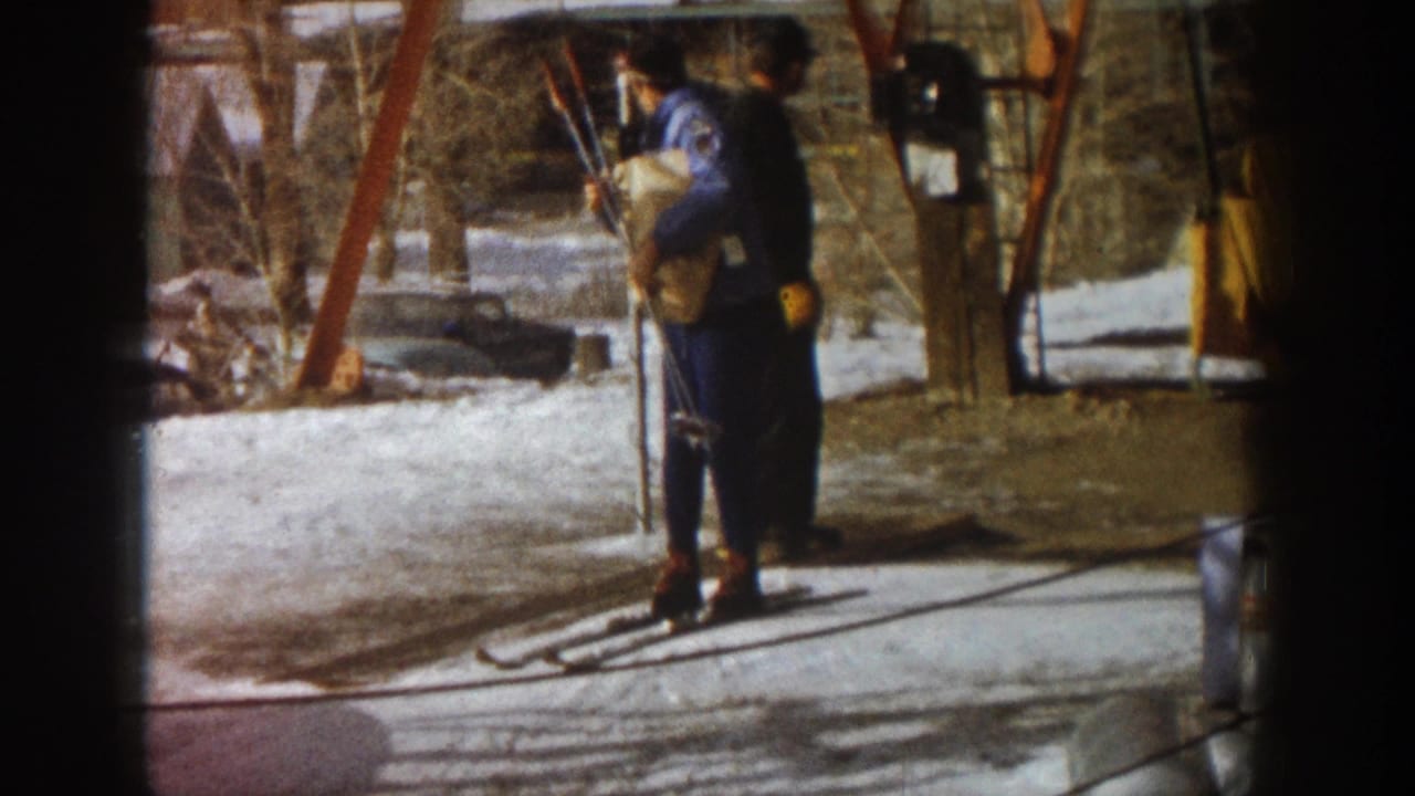 Two figures in dark blue denim ski clothing stand beside a painted metal lift structure at a ski area, consistent with Aspen, Colorado, circa 1961.