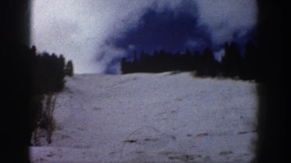 Wide shot of a snow slope and dark evergreen ridgeline under heavy cloud cover, continuing the scenic sweep from the preceding frame, consistent with Colorado mountain terrain, circa 1961.