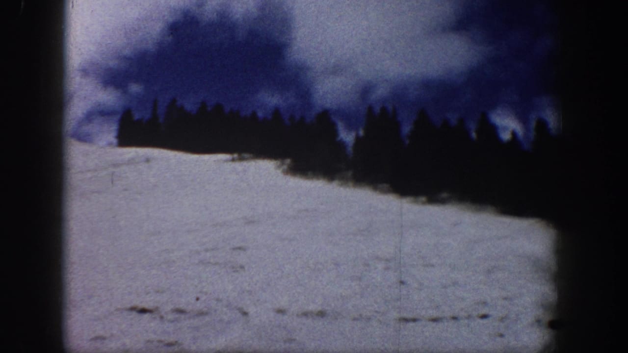 8mm color home movie frame showing a broad snow slope and a dense conifer treeline against a dramatic deep blue winter sky, consistent with Aspen, Colorado, circa 1961.