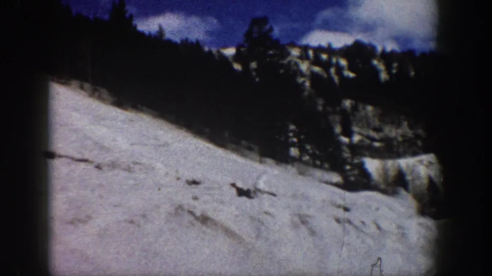 8mm home movie frame showing a snow slope, exposed rocky outcroppings, and snow-laden conifers under a blue winter sky, consistent with Aspen, Colorado, circa 1961.