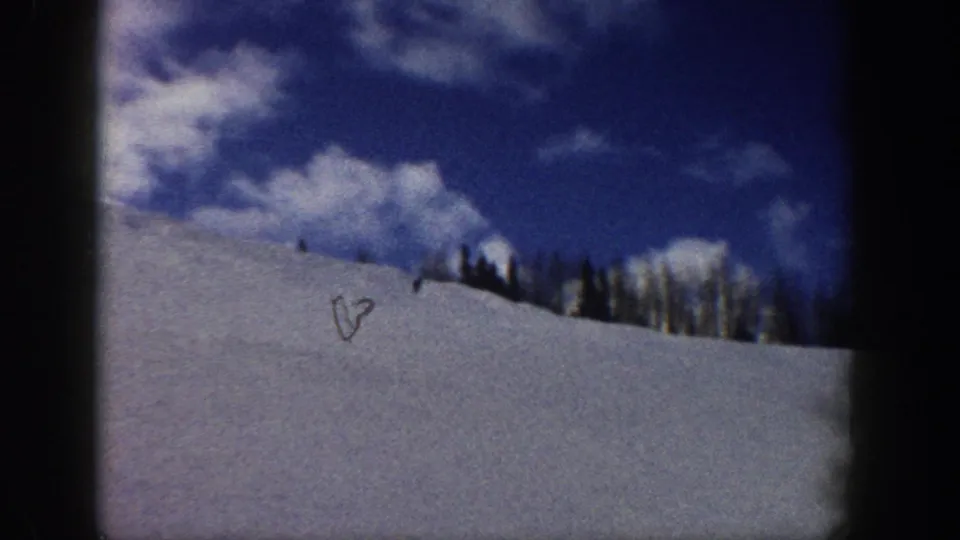 Wide 8mm shot with a tiny skier silhouette on a snowy slope under blue sky and cumulus clouds, likely Aspen, Colorado, circa 1961.