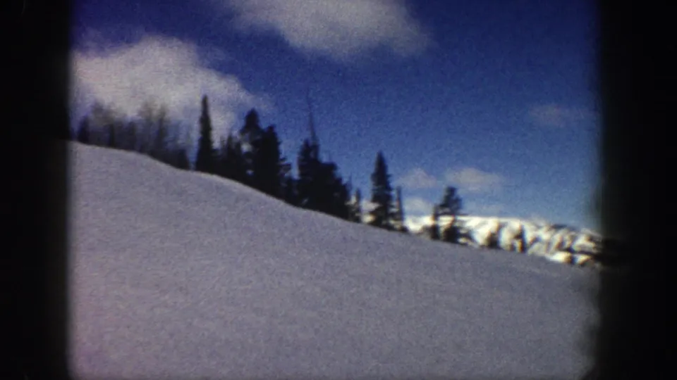 8mm home movie frame of a foreground snow slope, silhouetted conifers, and a distant snow-covered mountain ridge under blue sky, Aspen Colorado area, circa 1961.