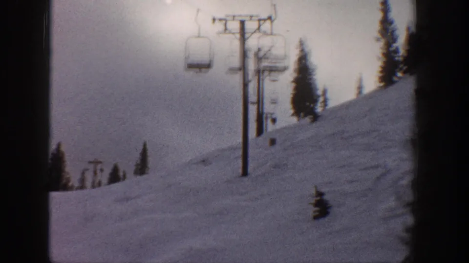 8mm home movie frame circa 1961 showing a broad snowy ski slope with chairlift ascending toward the tree line; bright sky upper left, possibly Aspen, Colorado.