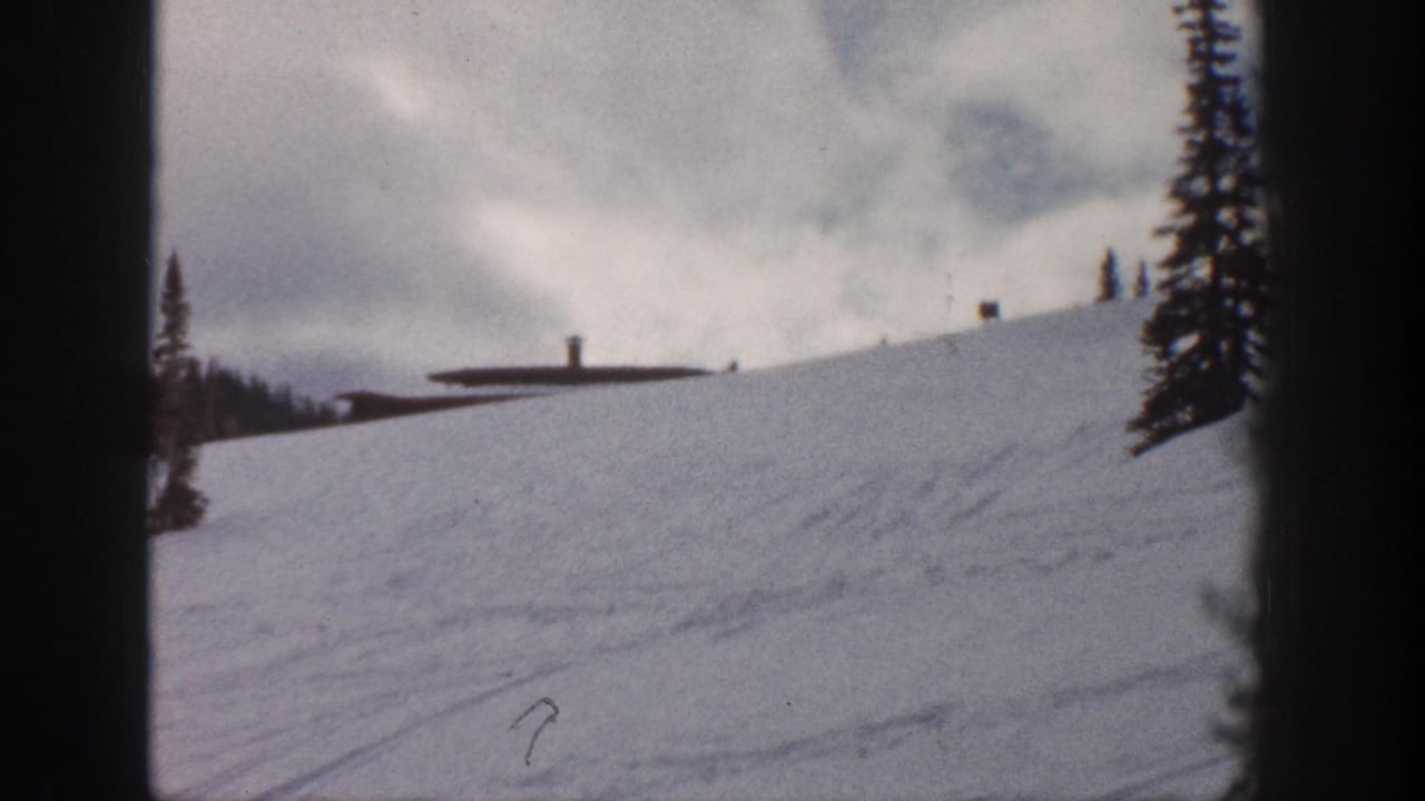 8mm frame of a snow slope at what may be Aspen, Colorado, circa 1961, showing a low ski station building and lift pole infrastructure against a heavily clouded sky.