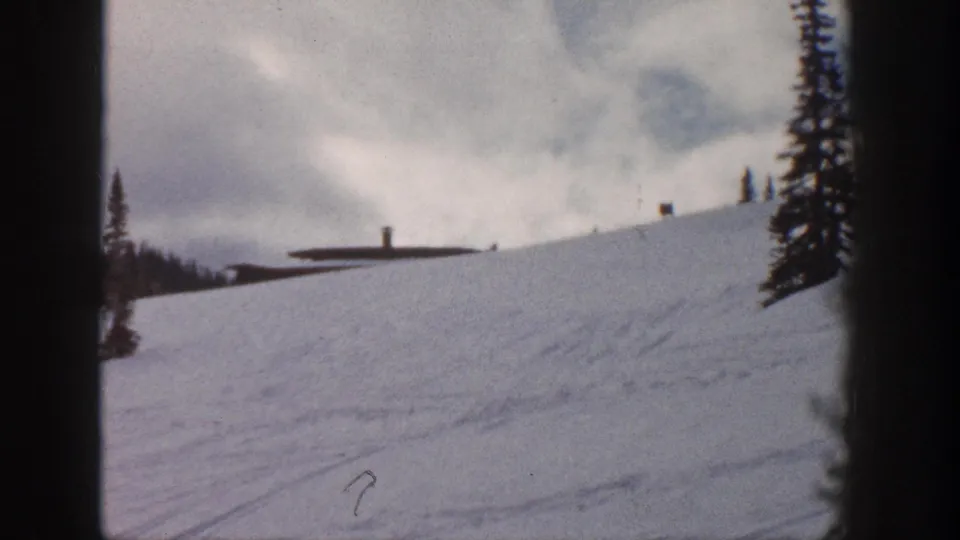 8mm frame of a snow slope at what may be Aspen, Colorado, circa 1961, showing a low ski station building and lift pole infrastructure against a heavily clouded sky.