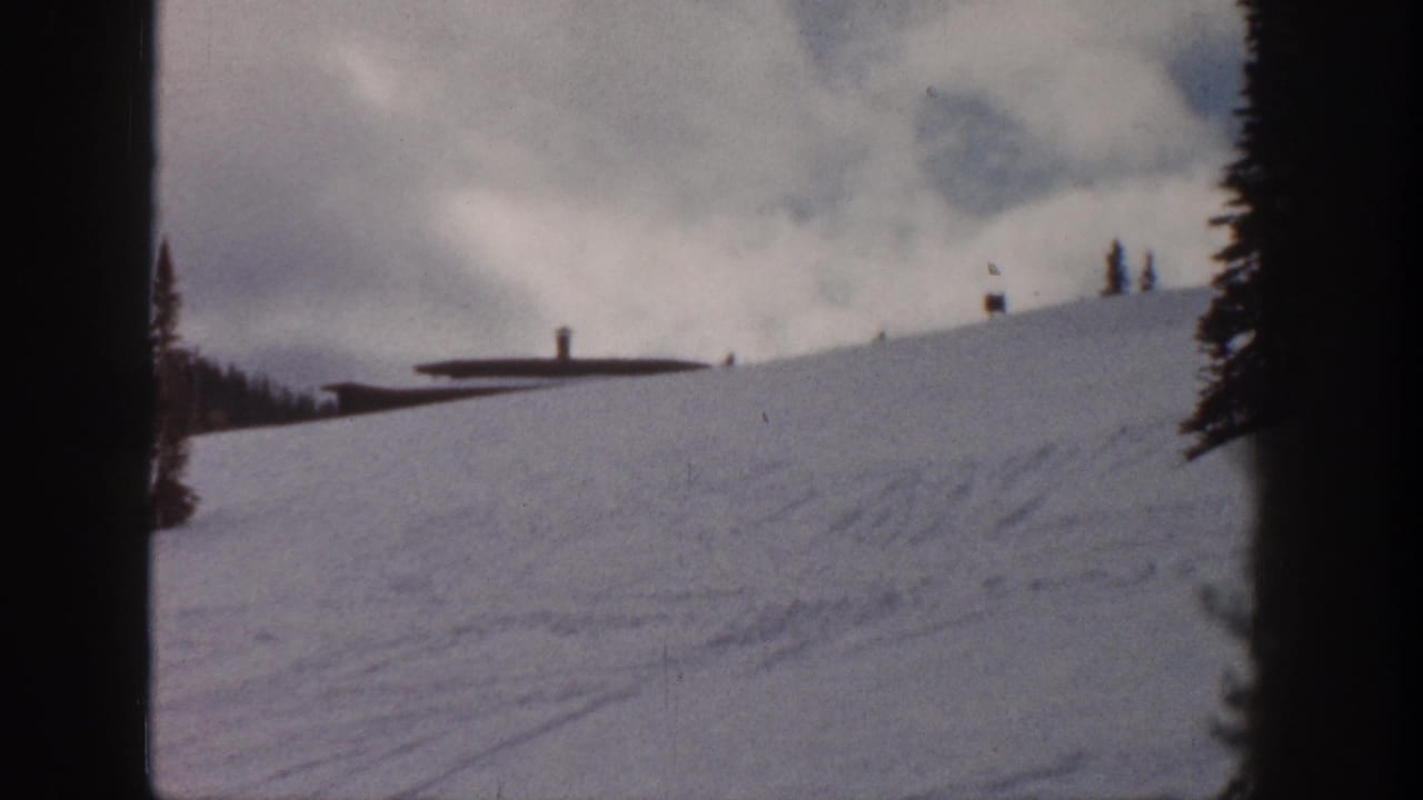 Snow slope with low dark-roofed building and lift towers near ridgeline; conifer trees at right edge and small marker or flag visible at upper ridge.