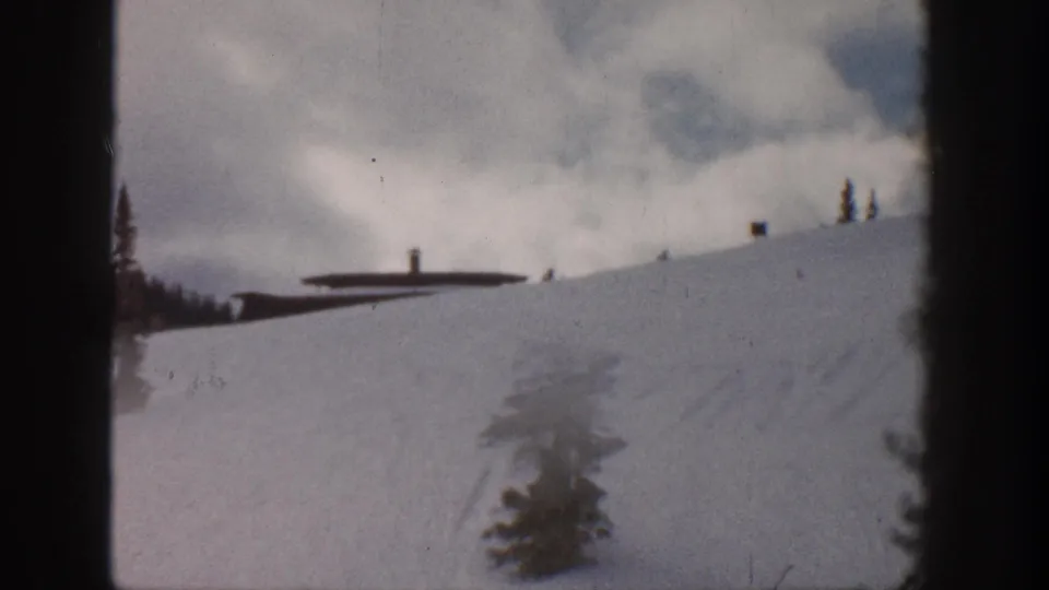 8mm frame of a snowy mountain slope, possibly Aspen, Colorado, 1961, with a foreground pine sapling, ski station building, and lift poles visible at the ridge.