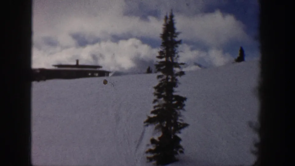 8mm home-movie frame from Aspen, Colorado, circa 1961 — evergreen tree in foreground, flat-roofed cabin on snowy slope, dramatic cumulus clouds above.