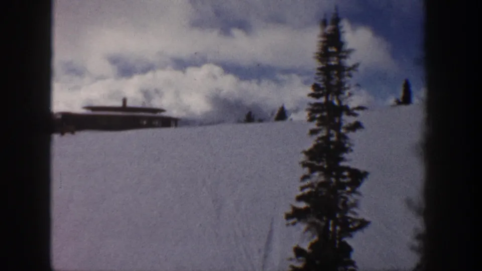 8mm pan frame from Aspen, Colorado, 1961 — cabin on snowy slope, evergreen tree moving right as camera pans, dramatic cloud formation above.