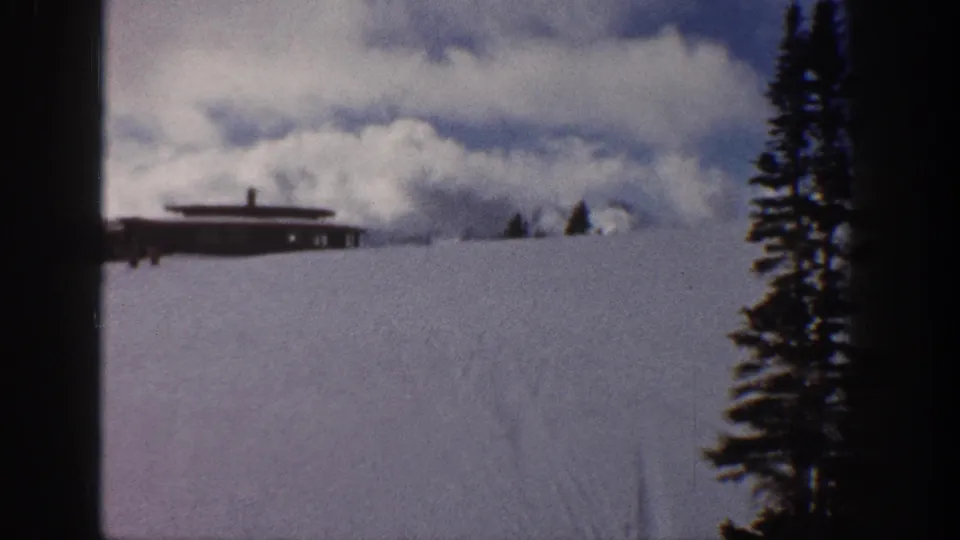 8mm pan frame from Aspen, Colorado, circa 1961 — cabin visible on snowy slope, second tall evergreen now central, hint of distant mountain peak, overcast sky.