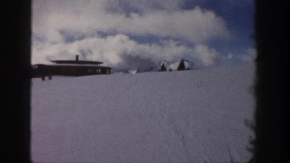 8mm pan endpoint from Aspen, Colorado, 1961 — cabin prominent on snowy slope, evergreen tree exiting right, cumulus clouds above open winter landscape.