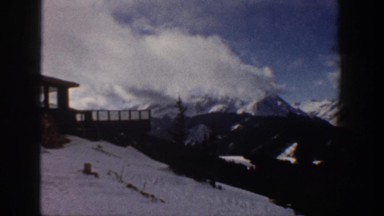 8mm home movie frame showing snow-covered peaks, a wooden railing deck at upper left, and a snowy slope; attributed to Aspen, Colorado, 1961.