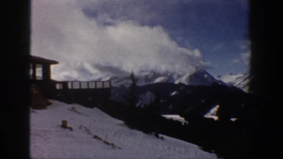 8mm home movie frame showing snow-covered peaks, a wooden railing deck at upper left, and a snowy slope; attributed to Aspen, Colorado, 1961.