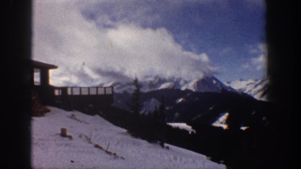 Snow-capped mountain range and wooden deck railing under a partly cloudy sky, 8mm film.