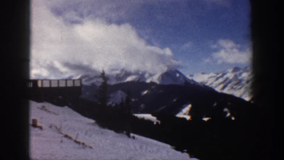 8mm home movie frame of a wide mountain panorama with wooden deck railing shifted to lower left as the camera pans; attributed to Aspen, Colorado, 1961.
