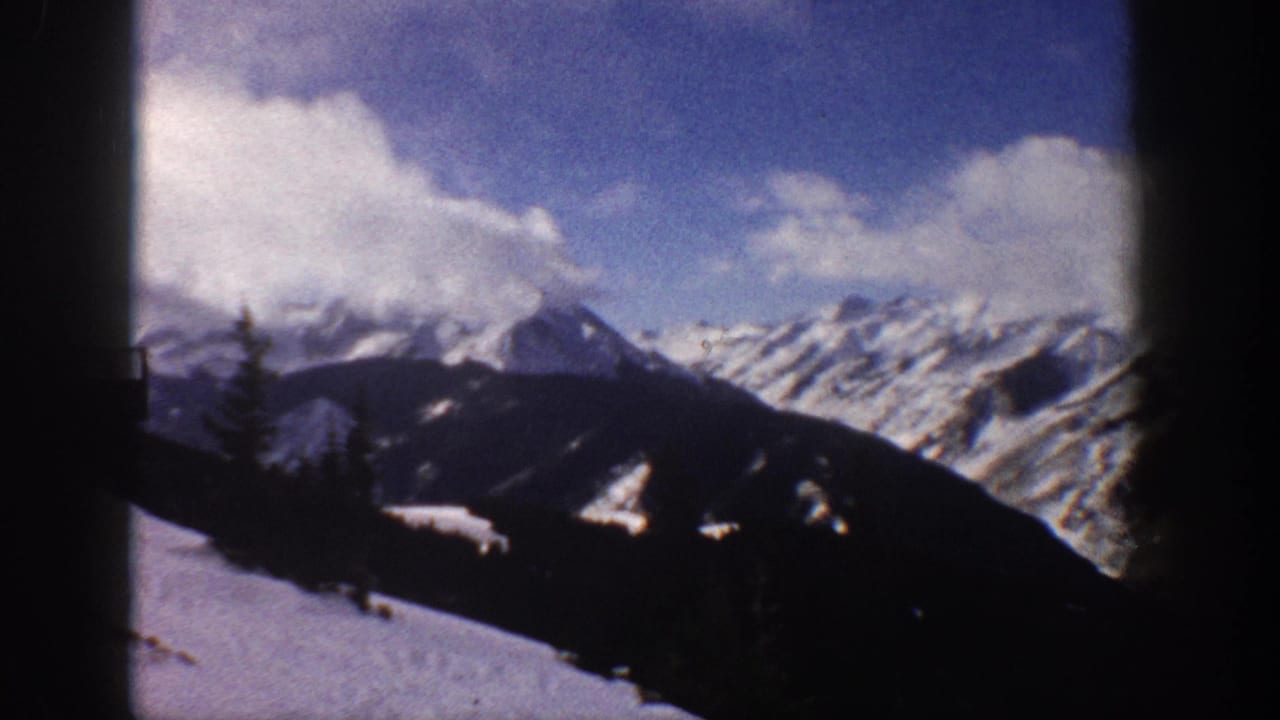 8mm home movie frame from Aspen, Colorado, 1961, showing snow-covered Rocky Mountain peaks and a dark foreground ridge under a blue sky with white clouds.