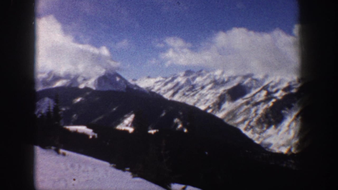 Slightly panned 8mm frame of snow-covered mountain ridge and peaks against blue sky with scattered clouds; dark vignetting at frame edges.