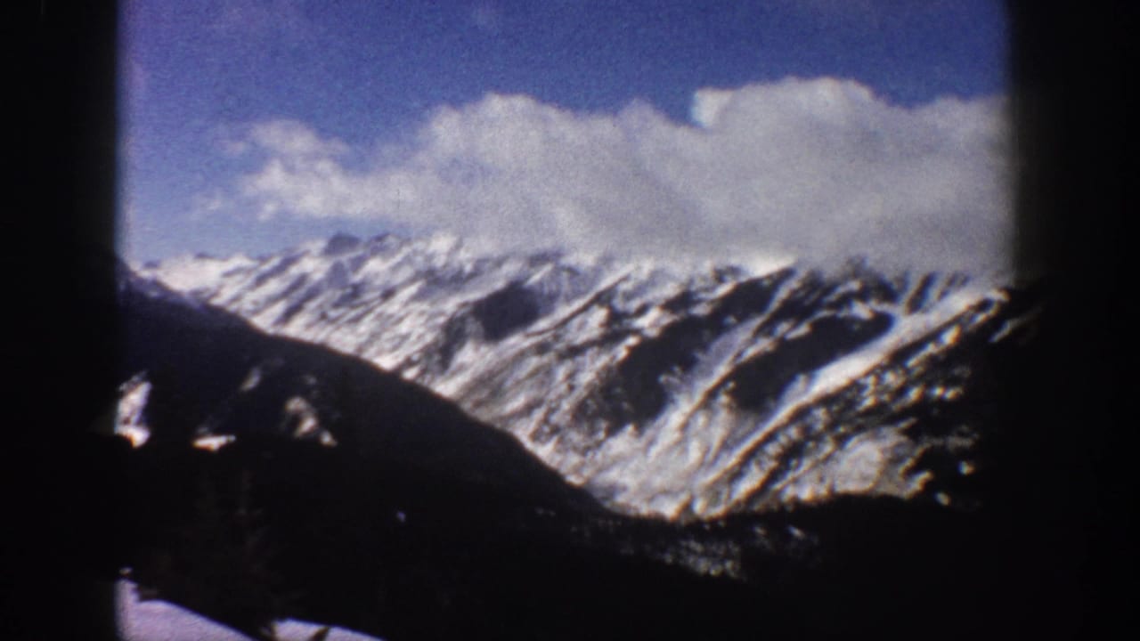 Snow-capped mountain range seen through a dark framing aperture, with deep blue sky and white cumulus clouds above a shadowed rocky ridgeline.