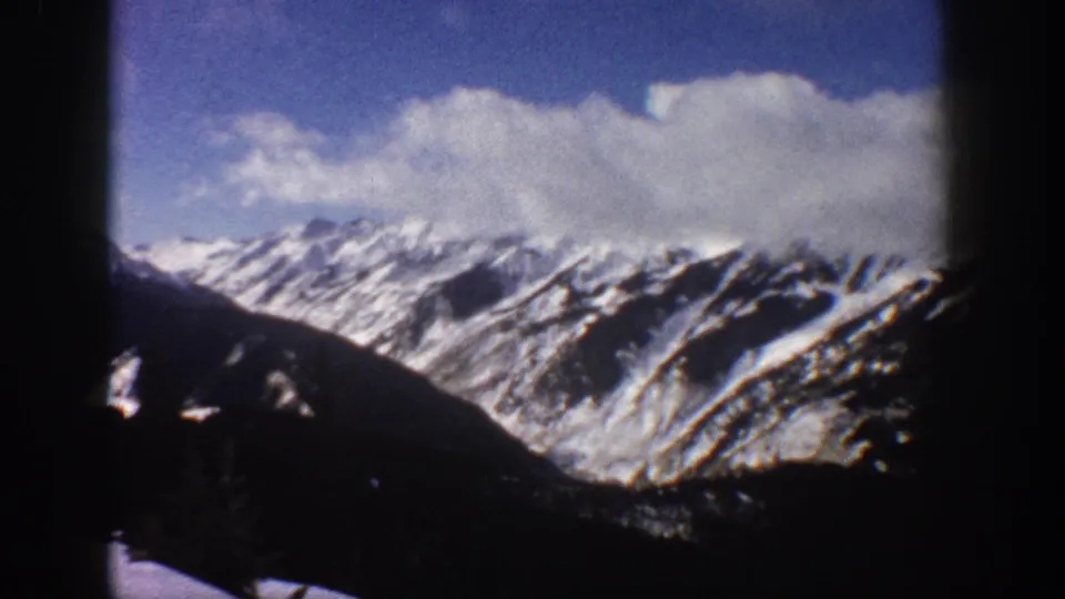 1961 8mm home movie frame of snow-covered mountains near Aspen, Colorado, framed through what appears to be a window or gondola opening, with cumulus clouds above the peaks.