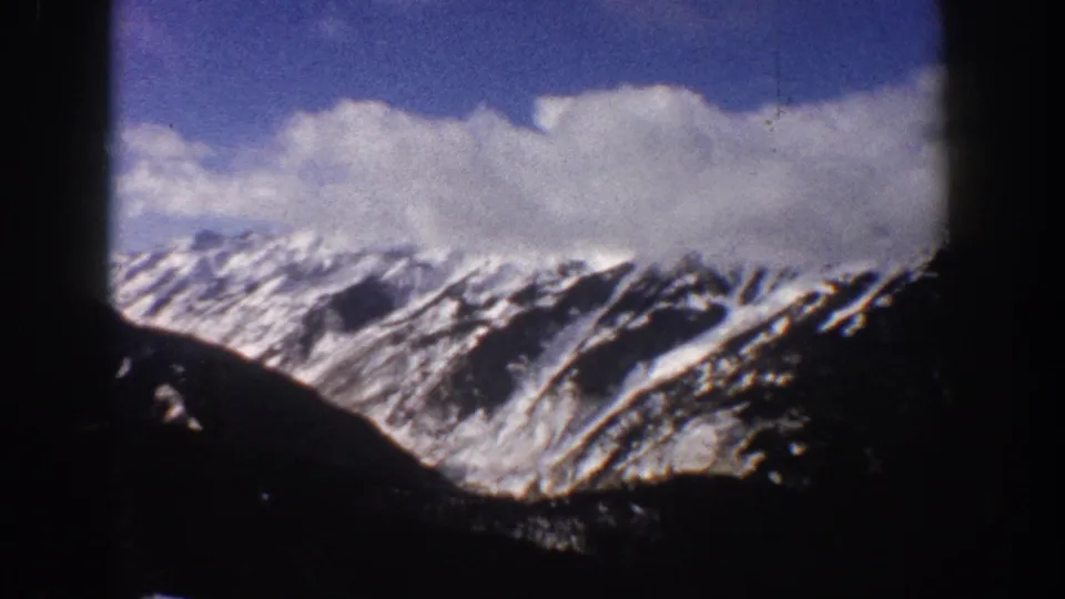 Continuation of an 8mm pan across snow-covered mountains near Aspen, Colorado, 1961, with blue sky and massed white clouds visible above the peaks and a dark foreground ridge.
