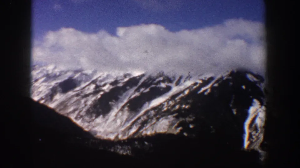 Mid-pan 8mm frame of snow-covered mountains near Aspen, Colorado, 1961, with what may be ski lift towers at the lower slope and large cumulus clouds above the peaks.