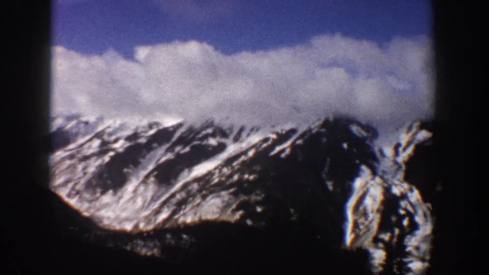 Final 8mm frame of snow-covered mountains near Aspen, Colorado, 1961, showing what may be ski lift towers at lower right, with large clouds above snow-laden peaks.