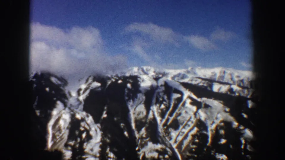 8mm film frame of the same Aspen, Colorado mountain face with snow-covered peaks, dark rocky ridges, and scattered clouds, circa 1961.