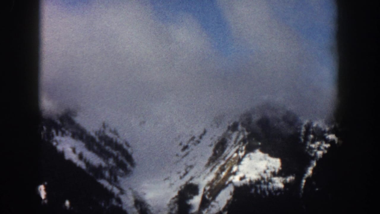 Snow-covered mountain peaks and rocky ridges with dark evergreen tree stands under a cloudy sky, 8mm film.