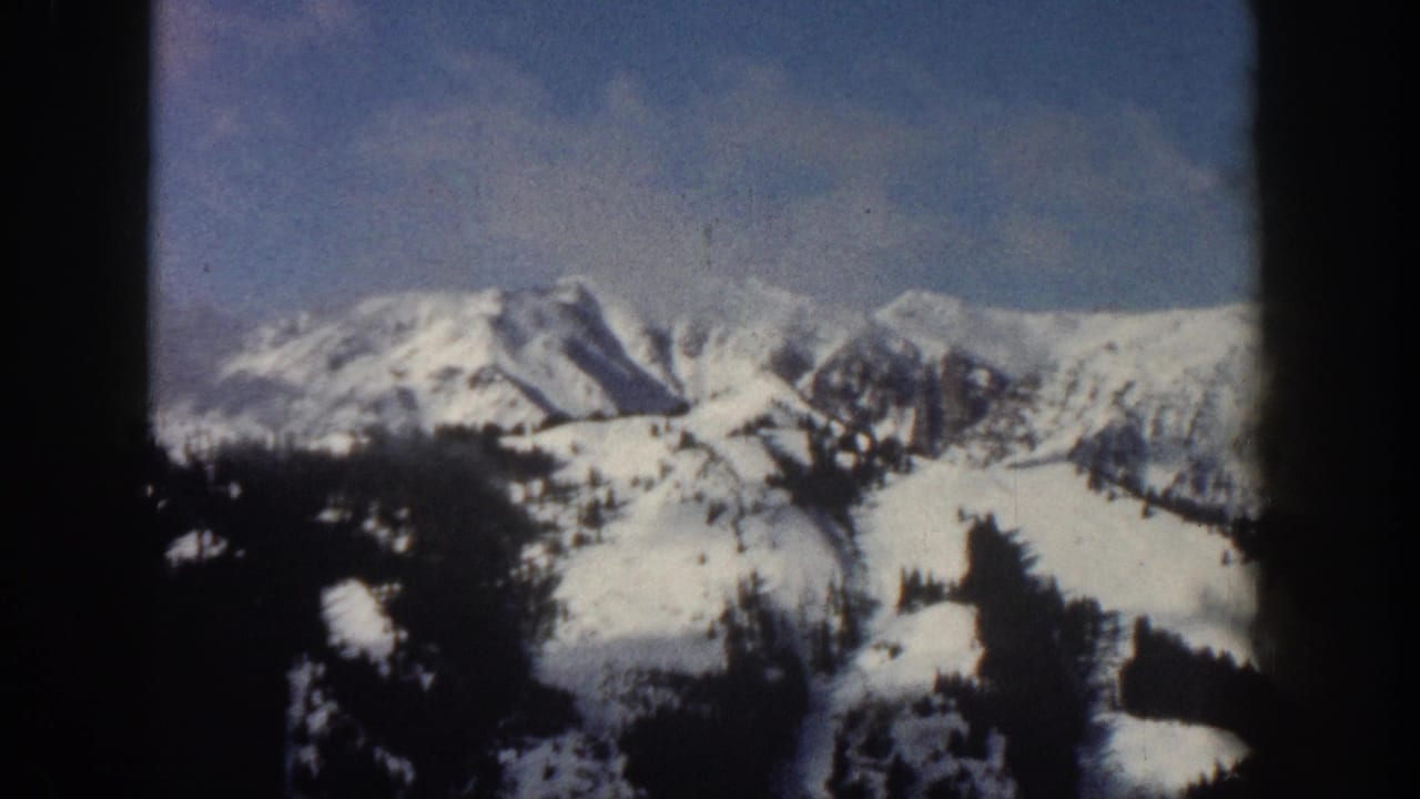 8mm home movie frame from Aspen, Colorado, 1961, showing snow-laden peaks and conifers under a pale blue winter sky with scattered clouds.